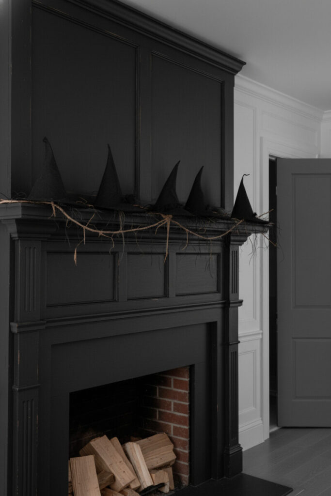 Clean black fireplace topped with witch hats, dried branches, and straw garland, a minimalist Halloween mantel decor display.