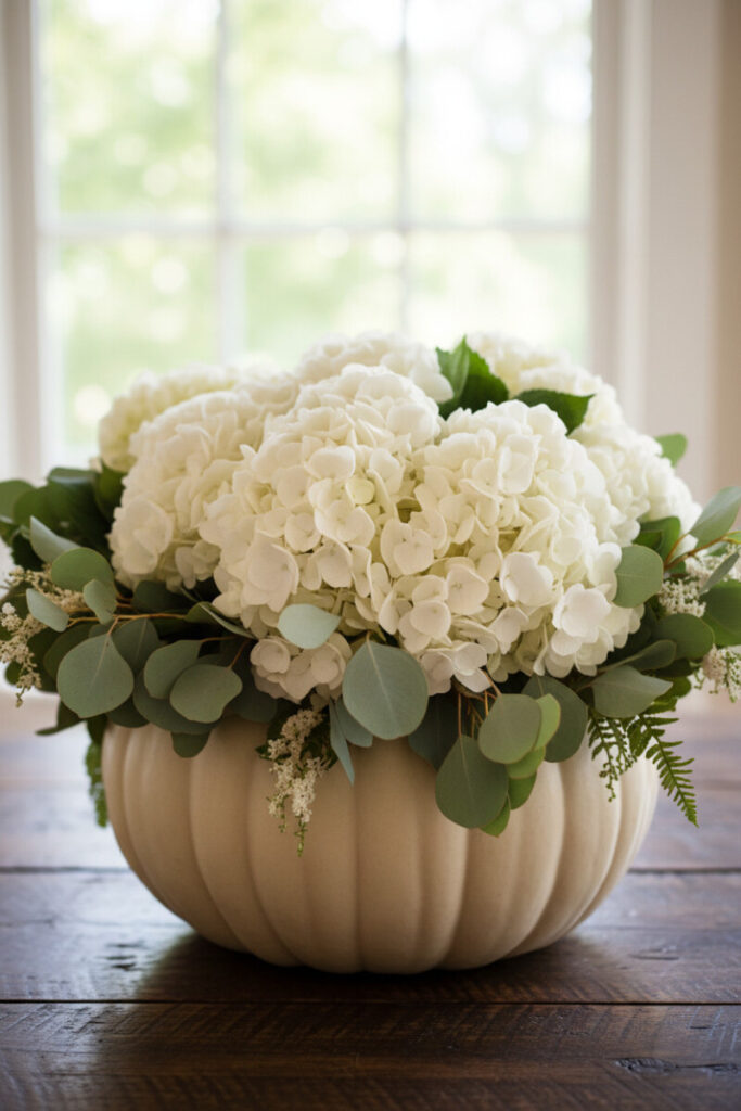 Cream pumpkin centerpiece filled with white hydrangeas and eucalyptus leaves, glowing with soft window light.