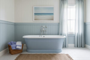 White walls over pale blue shiplap wainscot, coastal art, jute rug, and sheer blue curtains—breezy coastal blue bathroom style.
