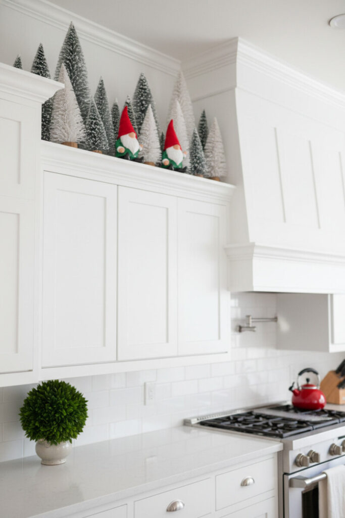 Playful elf vignettes nestled among flocked bottle-brush trees on top of white shaker cabinets—how to decorate kitchen cabinets for Christmas with whimsy.