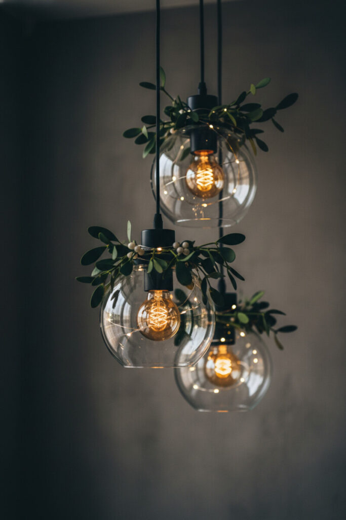 Cluster of clear glass pendant lights wrapped with mistletoe and micro LEDs—romantic contemporary holiday decor in moody chiaroscuro.