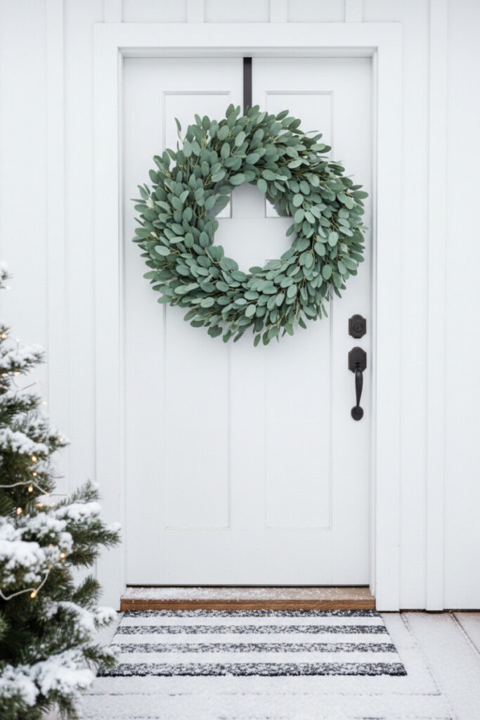Centered white door with full eucalyptus wreath and lightly snowy striped coir mat—serene scandinavian modern Christmas ideas for the entry.