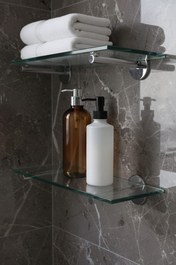 Two clear glass shelves with chrome brackets on dark veined marble, towels above and amber/white dispensers below.