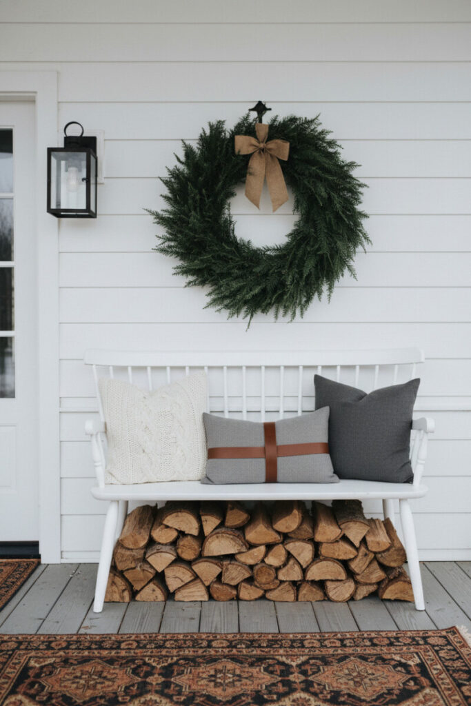 White bench over stacked firewood with evergreen wreath and cable-knit pillow—textural rustic winter front porch decorations look.
